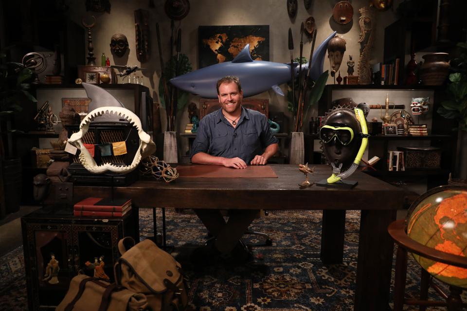 Josh Gates at a large wooden desk in a room filled with artifacts from around the globe.