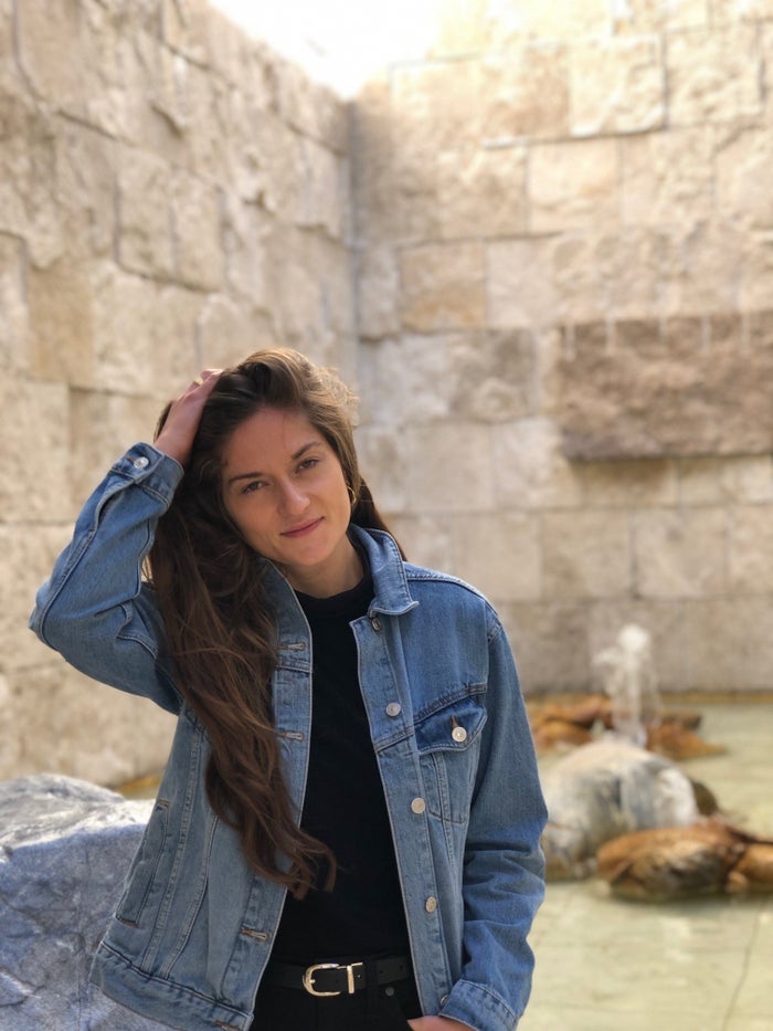 A woman in a jean jacket stands in front of brick walls, running her hand through her long hair